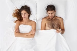 Attractive man feeling disappointed and depressed because of erectile dysfunction, looking at his penis under white cover, doesn't know what's the problem, angry wife lying next to him and waiting