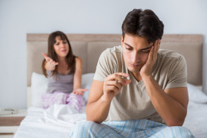 Man looks at erectile dysfunction treatment while partner is unhappy in bed behind him